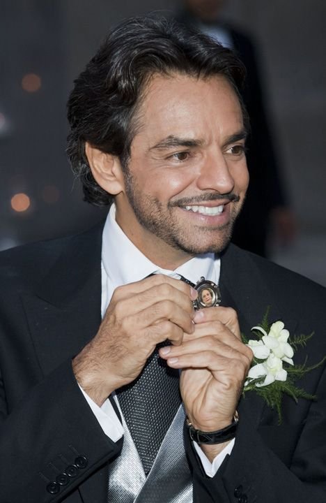 Eugenio Derbez in a tuxedo holding a locket with a photo, smiling.