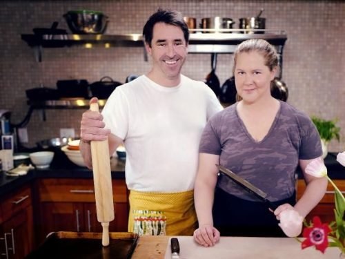 Chris Fischer with a rolling pin and Amy Schumer with a knife standing together in a professional kitchen.