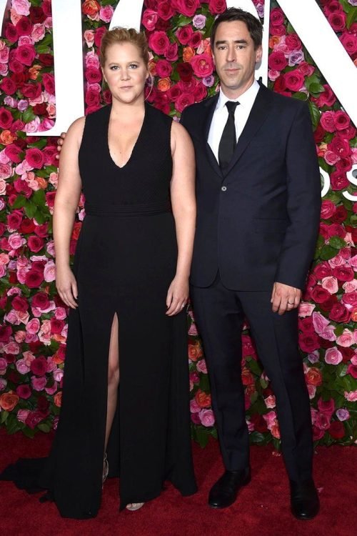 Chris Fischer in a dark suit and Amy Schumer in a black gown posing on a red carpet with a rose-covered backdrop.