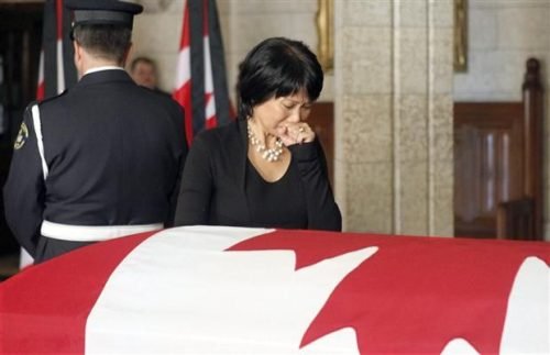 Olivia Chow standing solemnly beside a Canadian flag-draped casket, appearing emotional with her hand to her mouth.