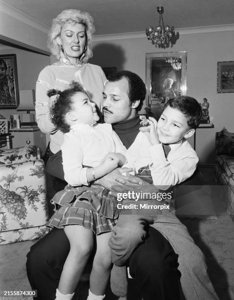 John Conteh holding his two children on his lap with his wife standing behind in a living room.