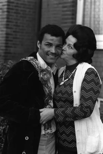 John Conteh with a ceremonial sash being kissed on the cheek by his mother outdoors.