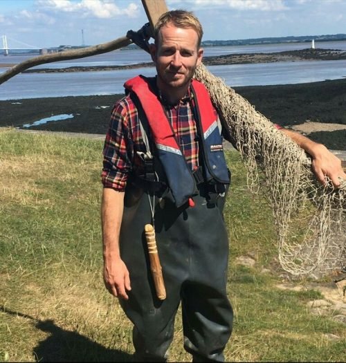 Jonnie Irwin in fishing gear holding a net near a coastal area.
