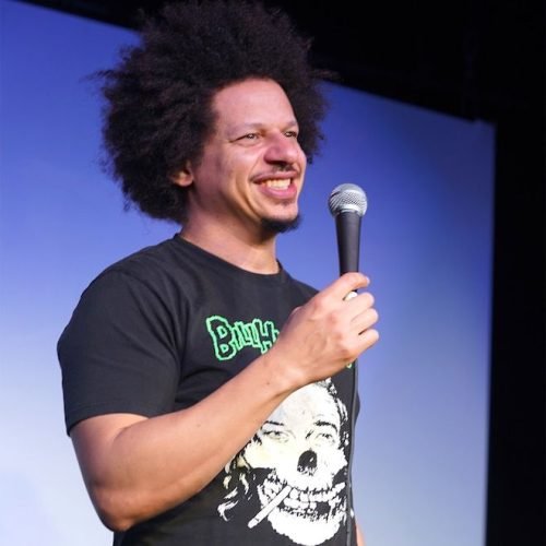 Eric Andre smiling while holding a microphone on stage during a stand-up comedy performance.