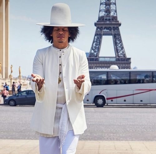 Eric Andre dressed in all white with a large wide-brimmed hat, standing with eyes closed and hands open in front of the Eiffel Tower.