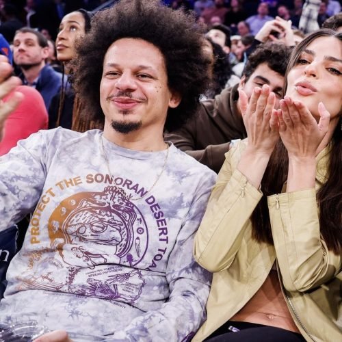 Eric Andre smiling while sitting courtside at a basketball game next to a woman blowing kisses toward the camera.