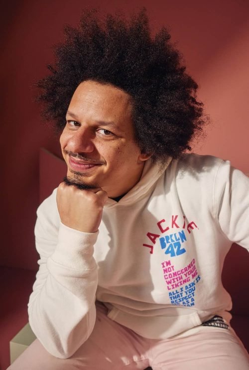 Eric Andre smiling and resting his chin on his fist while wearing a white hoodie with colorful text.