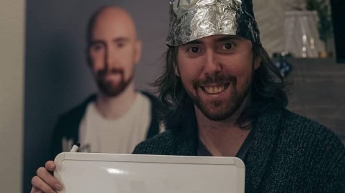 Asmongold while wearing a tin foil hat and holding a whiteboard, with a poster of a bald version of himself in the background.