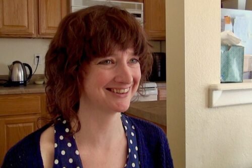Amber O Hearn with short, curly brown hair sits in a kitchen, wearing a blue polka-dot top and a dark cardigan.