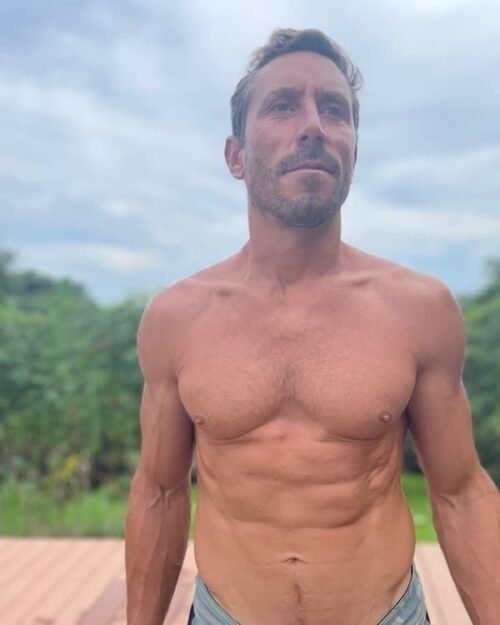 Paul Saladino stands outdoors with a focused expression, showing a muscular upper body against a blurred background of greenery and a cloudy sky.