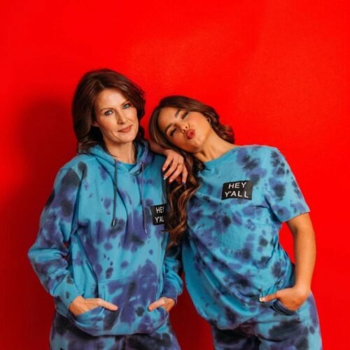Two women wearing matching blue tie-dye “HEY Y’ALL” outfits pose playfully in front of a bright red background. One stands with her hands in her hoodie pocket, while the other leans on her shoulder making a kissy face.