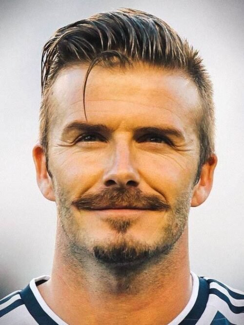 A close-up portrait of a smiling man with styled short hair, a full chevron mustache, and light stubble on his chin.