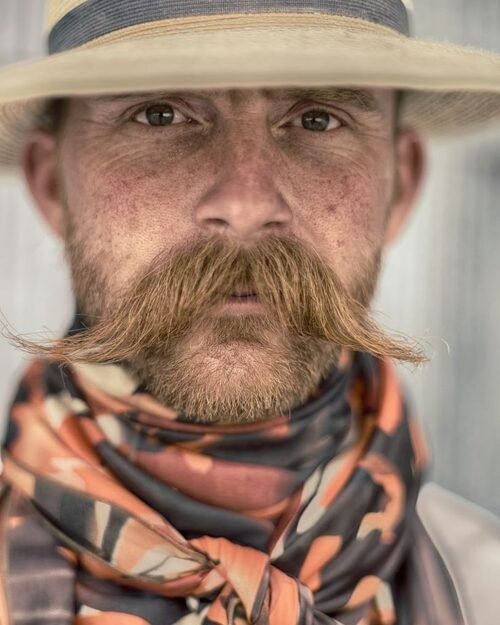 A man with a wide, curled cowboy-style mustache wearing a hat and a patterned scarf.