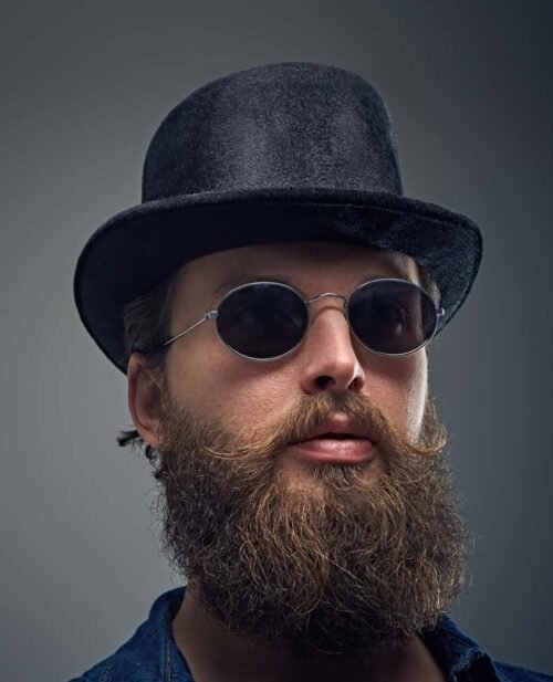 A bearded man wearing round sunglasses and a black top hat, sporting a full beard with curled mustache ends.