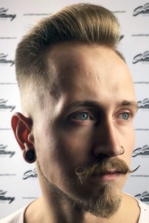 A man with a tall styled quiff, curled handlebar-style mustache, short beard, and ear gauges looking to the side.