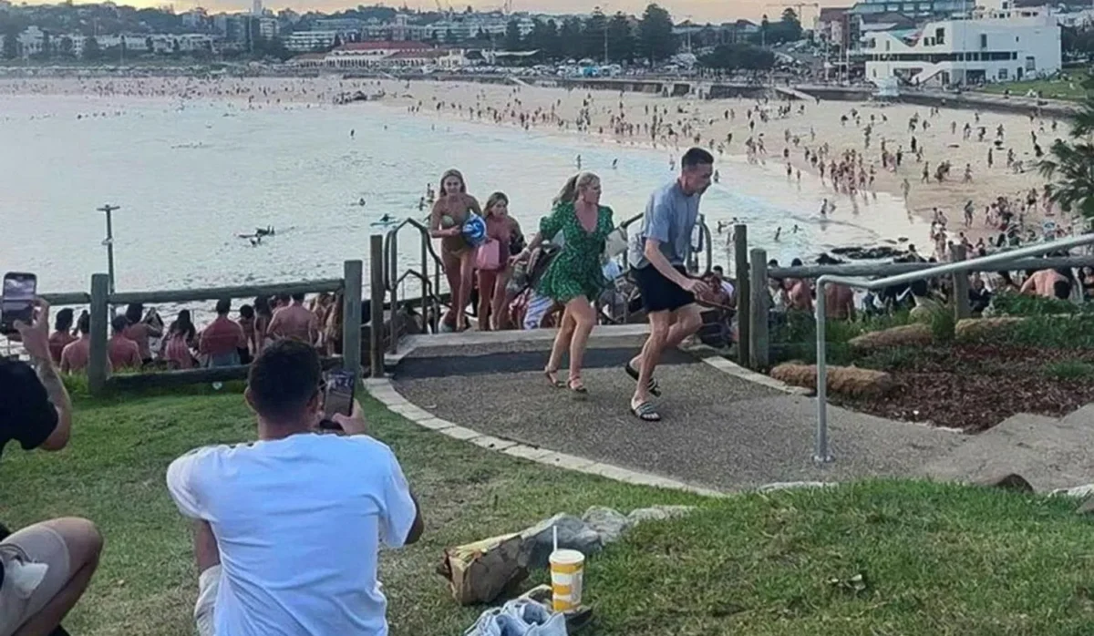 Crowded Bondi Beach in Sydney with people running up coastal steps as swimmers and beachgoers fill the shoreline during a busy summer day.
