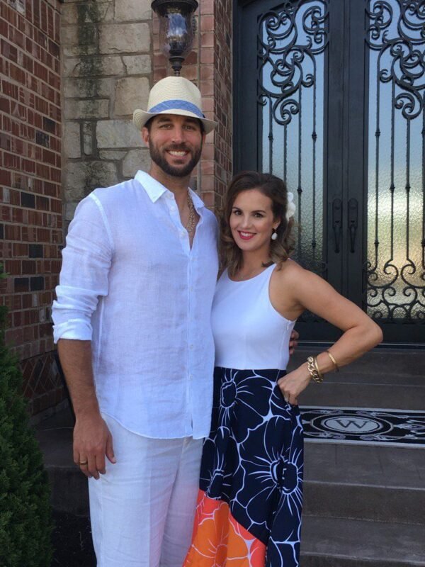 Smiling adam and his wife standing together outside a home entrance with decorative wrought-iron doors, dressed in stylish summer attire, posing for a casual outdoor photo.