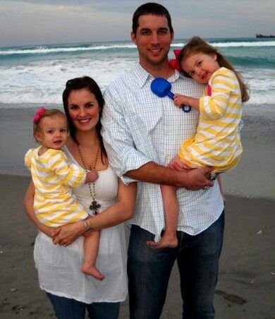 A family of four standing on a beach near the ocean, with two parents holding their young daughters while posing for a photo by the shoreline.
