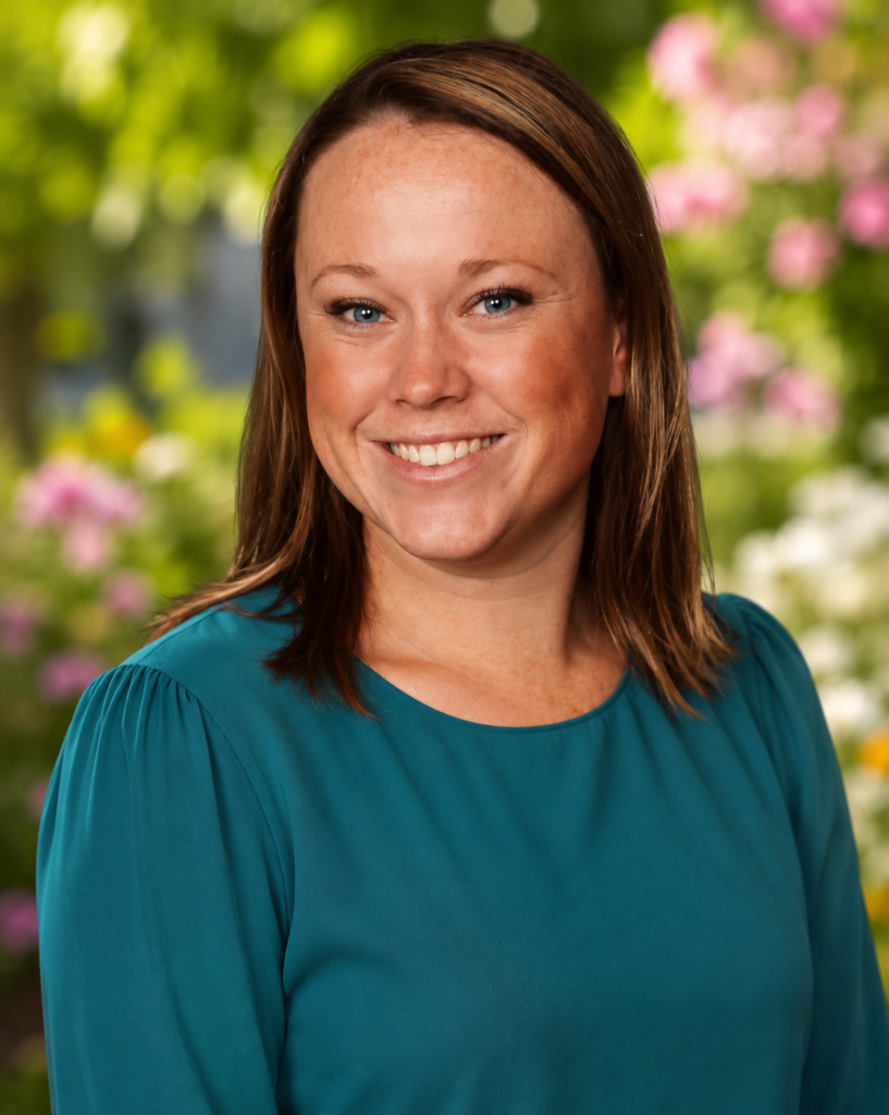 Smiling woman with brown hair wearing a teal blouse, standing in a colorful flower garden with a blurred natural background